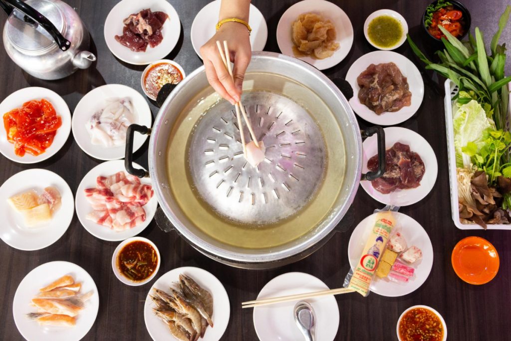 A person is holding chopsticks with a piece of lard over a hot mookata plate of food. at home